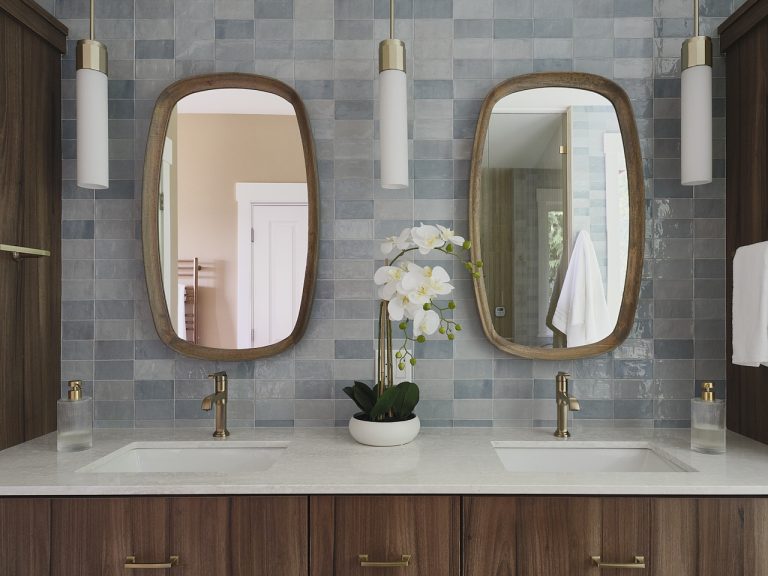 Double vanity with two brass faucets, blue tiled wall, and oval wooden-framed mirrors; white orchid centerpiece between sinks.
