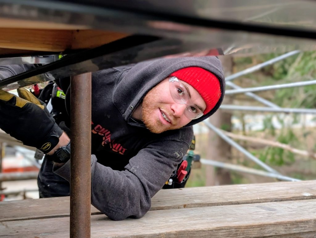 Man wearing safety goggles and red beanie leaning under a scaffold while holding a rusted vertical pipe during construction outdoors.