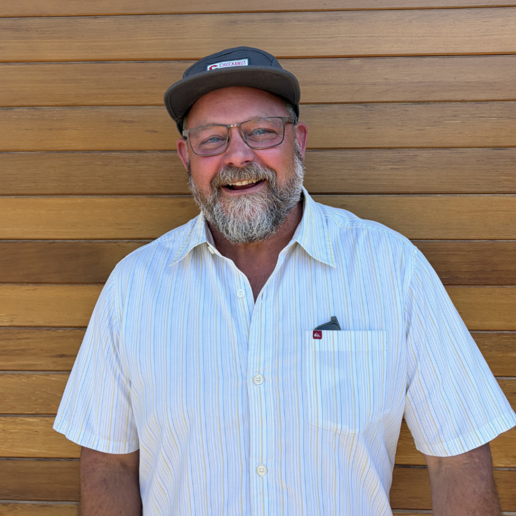 Smiling man with gray beard and glasses wearing a striped short-sleeve shirt and dark cap, standing against a wooden panel wall.