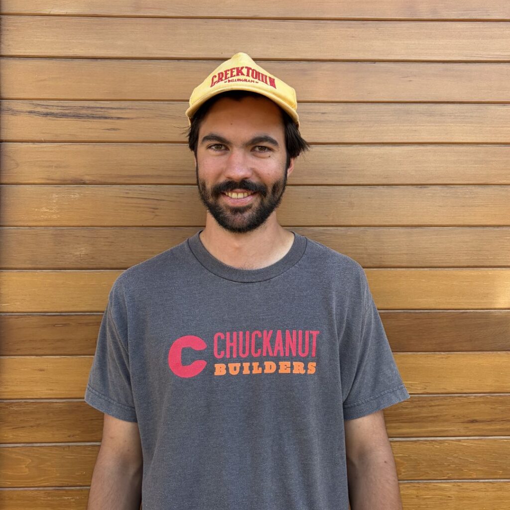 Smiling man wearing a yellow cap and dark gray Chuckanut Builders T-shirt stands against a wooden wall.