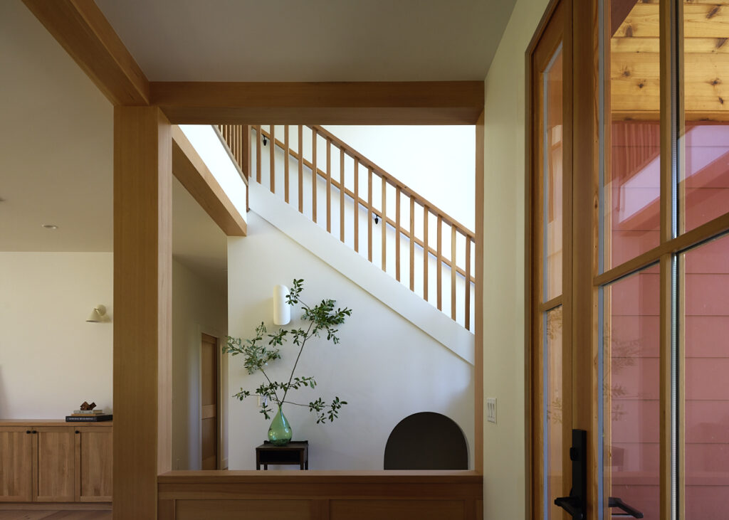Bright modern interior with exposed wooden beams, a diagonal staircase railing, and a glass door on the right; a small table holds a turquoise vase with greenery.